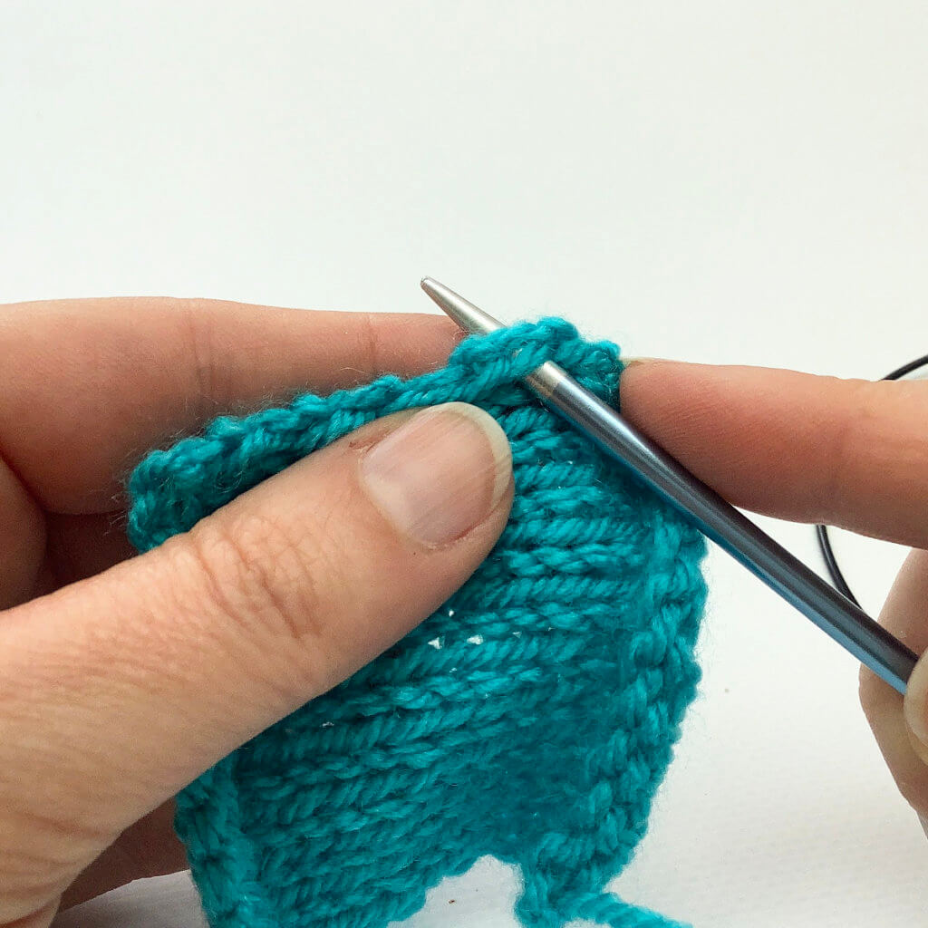 Picking up stitches; where to insert the knitting needle? Turquoise knitted swatch with a blue knitting needle inserted underneath both legs of a v-shaped stitch at the edge of the fabric.