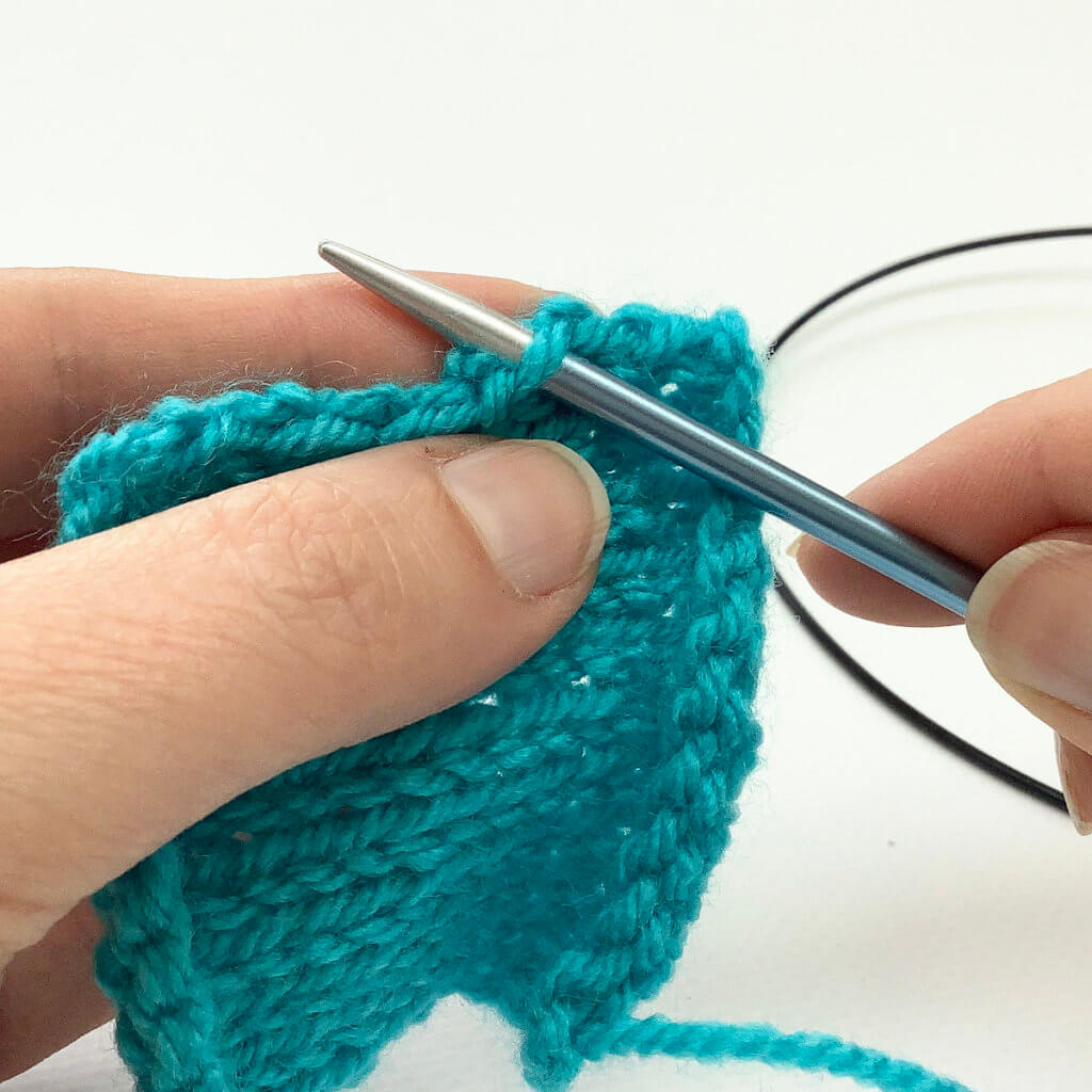 Picking up stitches: Turquoise knitted swatch with a blue knitting needle inserted underneath the left leg of a v-shaped stitch at the edge of the fabric.