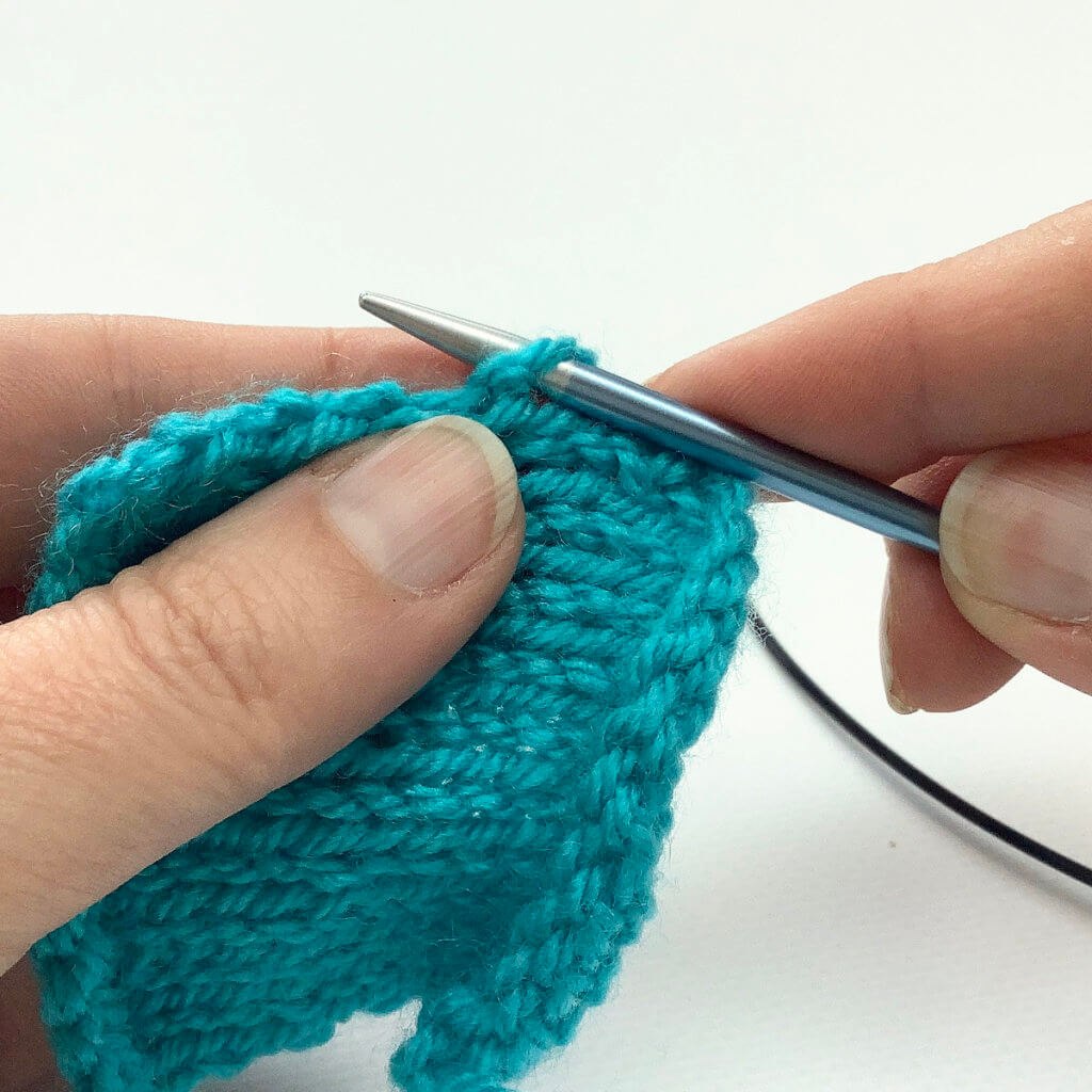 Picking up stitches: Turquoise knitted swatch with a blue knitting needle inserted underneath the right leg of a v-shaped stitch at the edge of the fabric.