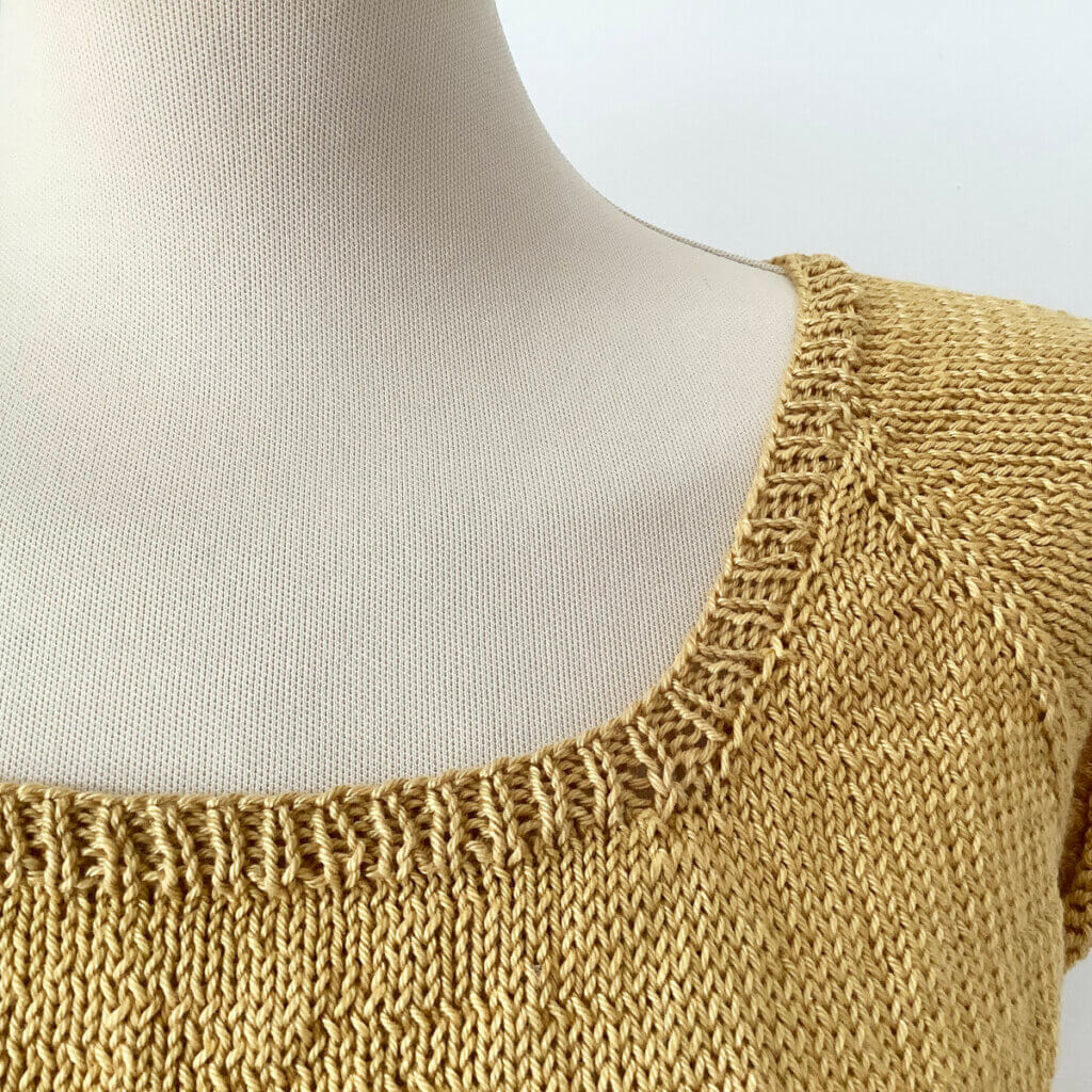 Yoke detail of a knitted tee in yellow yarn, with a finished neckband, shown on a mannequin.