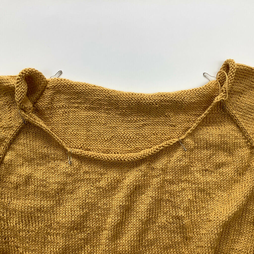 The yoke of an unfinished knitted tee in yellow yarn, now with bulb pins added to the transition points of the various neckline sections.