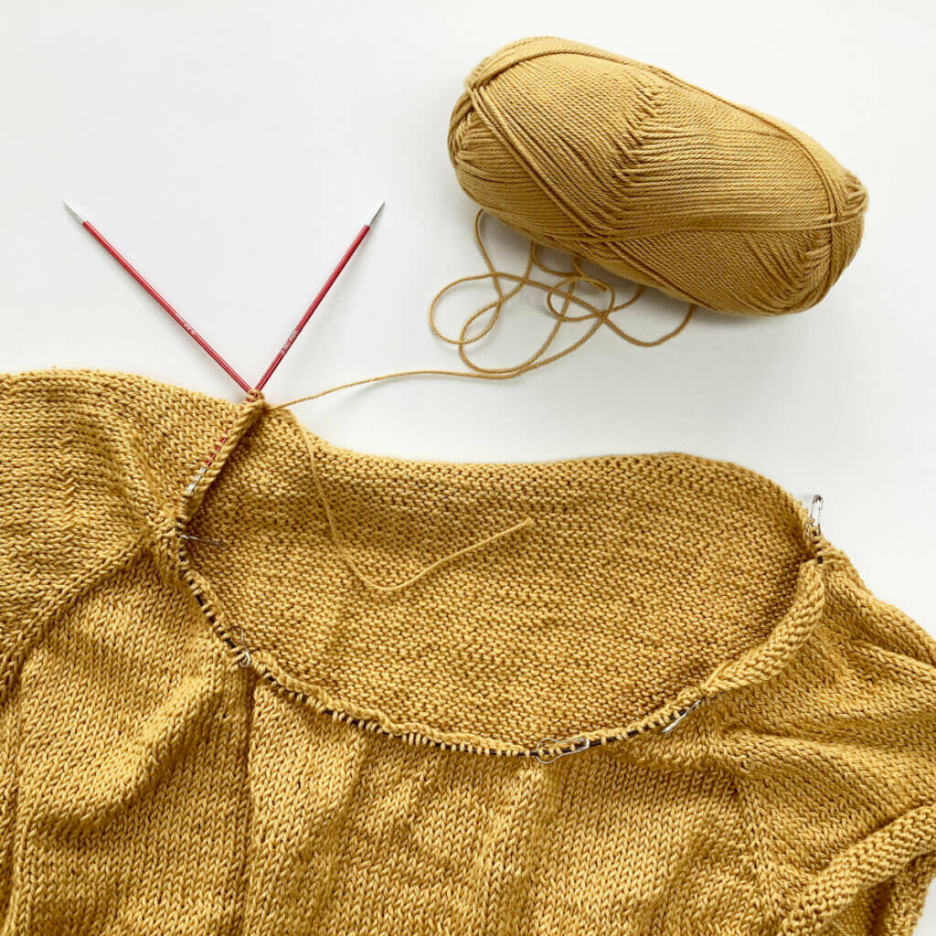 The yoke of an unfinished knitted tee in yellow yarn, with all neckline stitches on a circular needle.