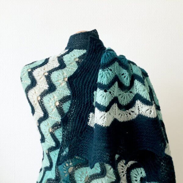 Close-up of a shawl in various teals, forming a gradient. It has wavy lace patterning, and a border in garter stitch. It appears draped over a mannequin, against a white background.