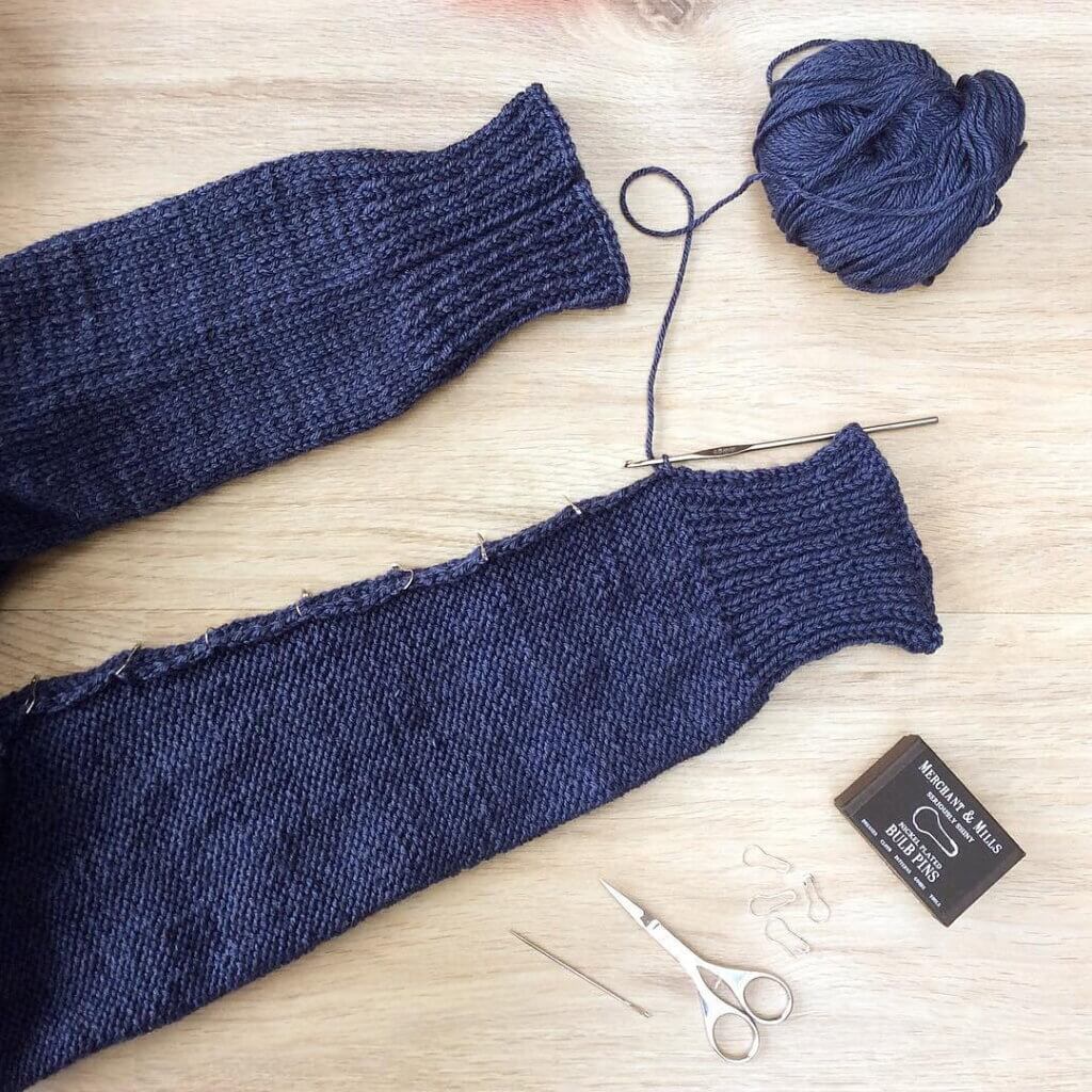 Flat lay view of the sleeves of a blue garment being seamed together using the crochet method. Bulb pins are being used to align the edges.